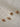 Four gold chain necklaces with ornate floral halo pendants and rectangular gemstones in pink, peach, and brown tones, arranged diagonally on a light beige textured surface.
